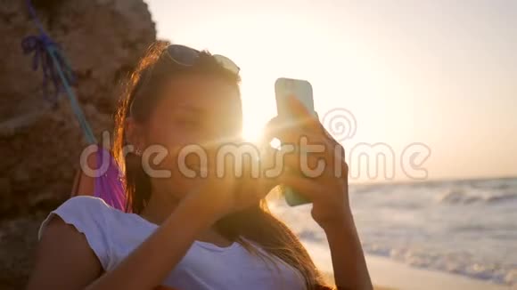 在海边的沙滩上放松的混血年轻女子在吊床上使用手机迎接令人叹为观止的金色日落视频的预览图
