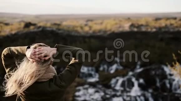 后景年轻的金发女人站在悬崖上看着冰岛的Barnafoss瀑布摸着头发视频的预览图