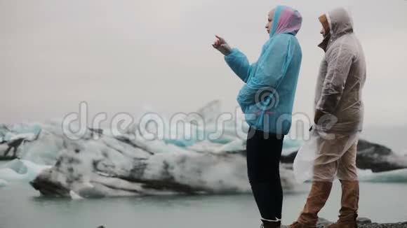 年轻的旅行夫妇穿着雨衣在冰岛的冰湖欣赏风景在寒冷的日子里大自然的游客视频的预览图
