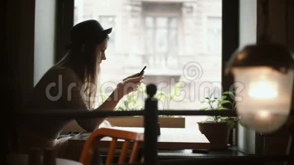 漂亮的年轻女人坐在靠近窗户的桌子旁用智能手机在社交网络中聊天视频的预览图