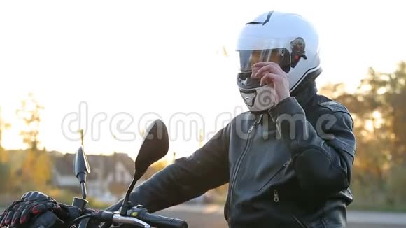 一个穿黑色皮夹克的年轻人坐在摩托车上在秋日日落前准备好白色头盔视频的预览图