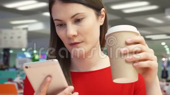 在食品场的快餐店里一个口渴的女孩从纸杯里喝咖啡使用手机视频的预览图