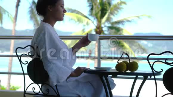 年轻快乐的女人穿着浴袍坐在阳台上喝咖啡在令人惊叹的海景上使用手机视频的预览图