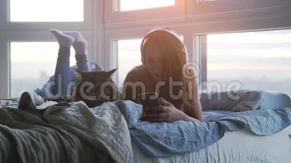 戴着耳机的年轻美女和可爱的缅因州浣熊猫一起放松听躺在床边的音乐视频的预览图