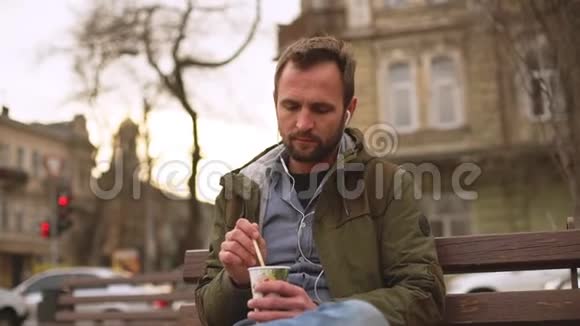 帅哥穿着牛仔裤衬衫在咖啡上画画棍子坐在长凳上听耳机里的音乐视频的预览图