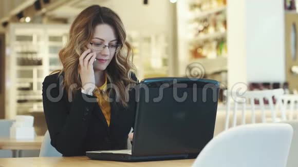 一位年轻的女商人在咖啡馆工作接电话穿西装在笔记本电脑上工作的女孩视频的预览图