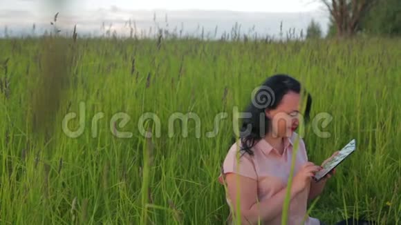 微笑的女人坐在绿草中在公园里的平板电脑上工作视频的预览图