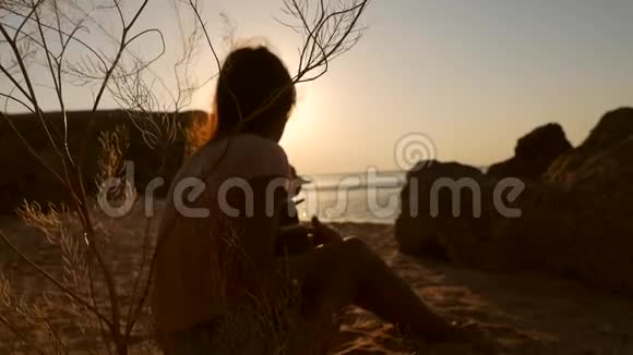 年轻的嬉皮士波霍女孩坐在沙滩上弹着四弦琴吉他在令人惊叹的日落附近的海洋高清慢镜头视频的预览图