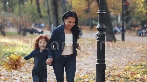 快乐的年轻母亲和她的小红发女儿一起在一个秋天的公园里跑步他们笑着玩视频的预览图