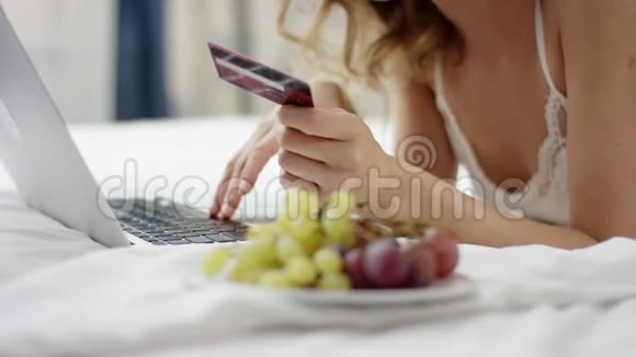 穿着睡衣的迷人女人躺在床上用她的笔记本电脑和信用卡视频的预览图