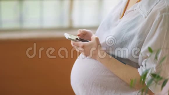 站在木窗前的年轻女性穿着白色t恤轻敲智能手机的孕妇侧视视频的预览图