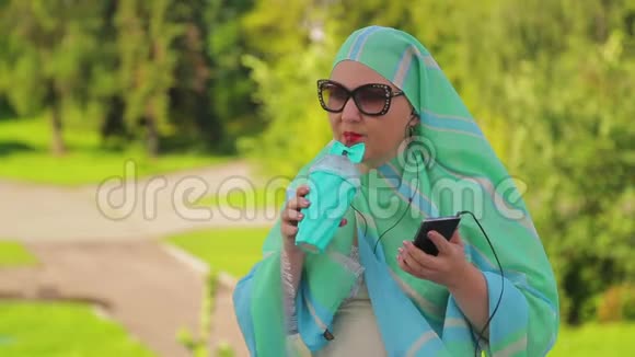 公园里一位戴着浅色围巾的年轻穆斯林妇女正在喝咖啡带着耳机在信使里说话视频的预览图