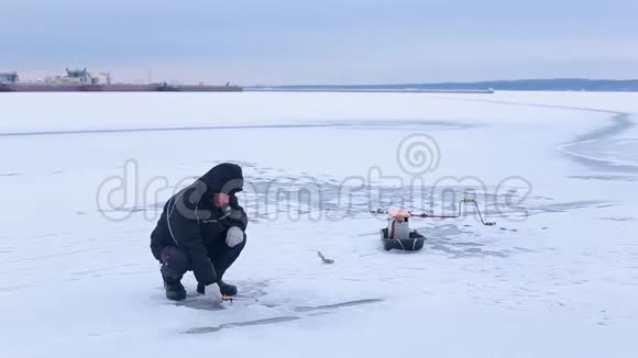 穿着深色衣服的老渔夫在河港的背景下在冰冻的河面上用冬季的鱼竿钓鱼视频的预览图