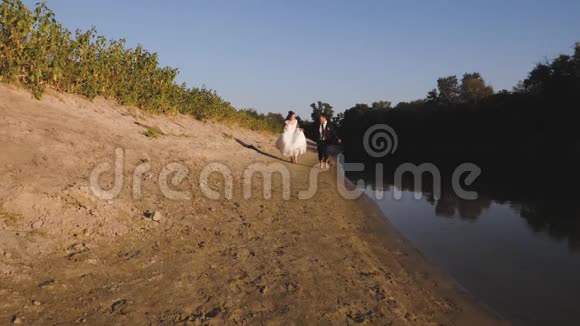幸福的新娘和新郎赤脚在河边的海滩上奔跑视频的预览图