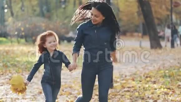 快乐的年轻母亲和她的小红发女儿一起在一个秋天的公园里跑步他们笑着玩视频的预览图