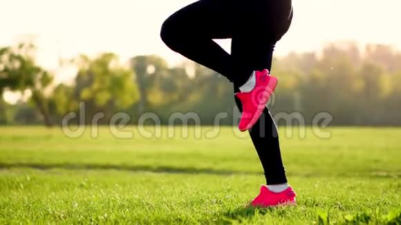 日落时分在草地上一个运动的女孩穿着白色的上衣和粉红色的运动鞋在提起前进行热身运动视频的预览图