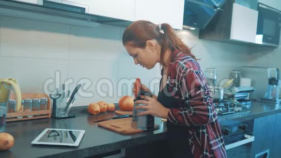 那个女孩把胡萝卜揉在数字平板电脑上的烤盘上美丽的女人在烹饪和测试食物视频的预览图