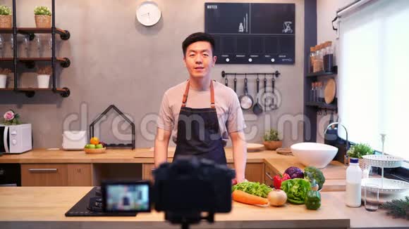 年轻的亚洲男人在厨房用相机录制视频微笑的亚洲男人在食品博客概念与水果和蔬菜视频的预览图