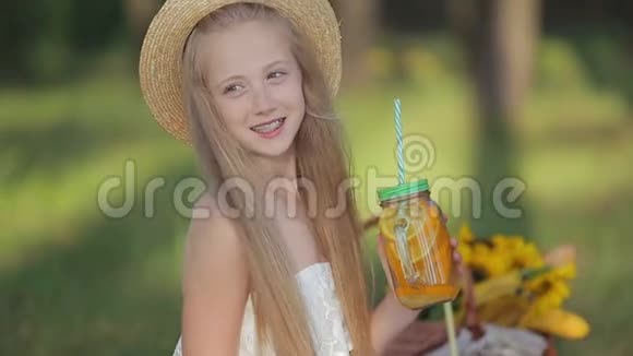 年轻女孩穿着浪漫的白色连衣裙和帽子坐在野餐毯上喝鸡尾酒视频的预览图