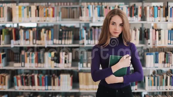 微笑的女学生拿着一本书站在图书馆的书架前视频的预览图