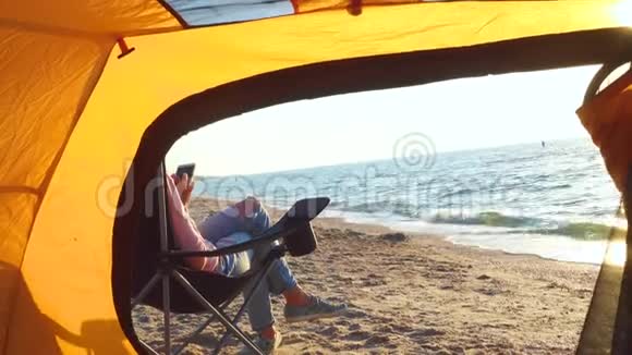 在沙滩上靠近徒步帐篷的椅子上放松的女孩视频的预览图