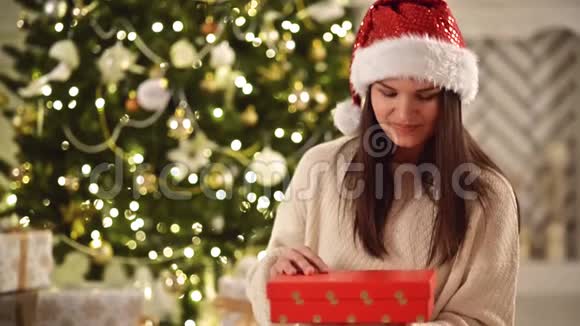 圣诞帽圣诞礼物盒和微笑的女人在圣诞树背景有着长长的黑发和圣诞老人的美丽的布鲁内特视频的预览图