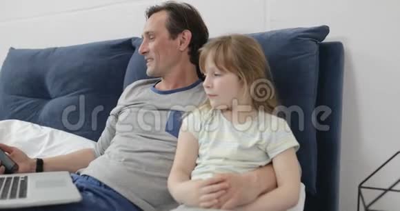 父亲和女儿用笔记本电脑快乐地微笑着母亲拿着数字平板躺在床上的家庭视频的预览图