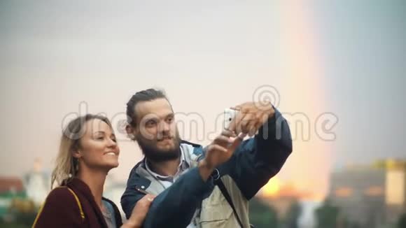 幸福夫妇站在市中心日落在智能手机的背景上拍摄带有彩虹的自拍照片视频的预览图