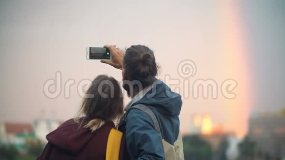 年轻漂亮夫妇在智能手机上拍摄市中心景观日落和彩虹的背面景色视频的预览图