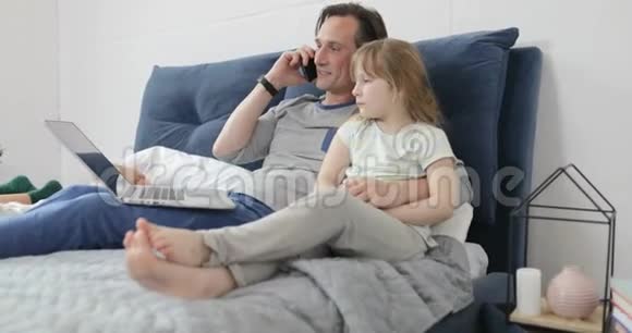 父亲拥抱女儿谈电话一起坐在床上在卧室里用笔记本电脑视频的预览图