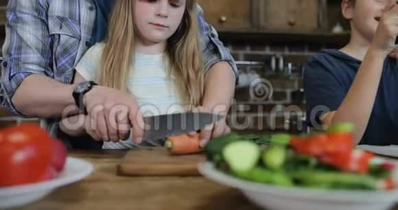 父亲帮助女儿切蔬菜而儿子和母亲在厨房使用手机父母有两个孩子视频的预览图