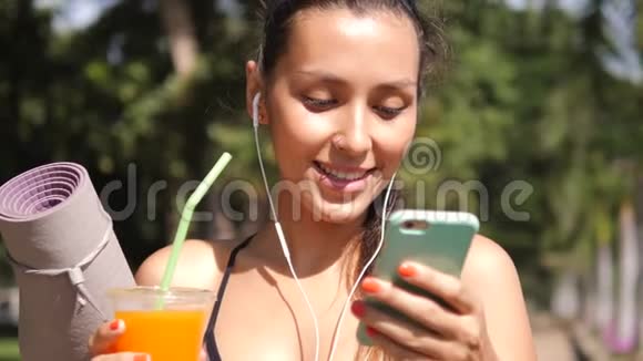吸引人的健身妇女使用手机应用程序和饮用新鲜橙汁后上午瑜伽练习在城市公园视频的预览图