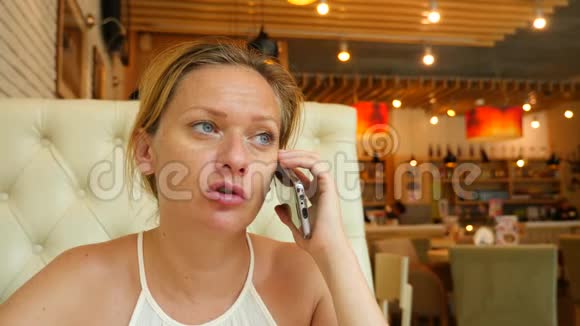 在炎热的夏天金发女郎坐在咖啡馆里微笑着打电话4k慢动作视频的预览图