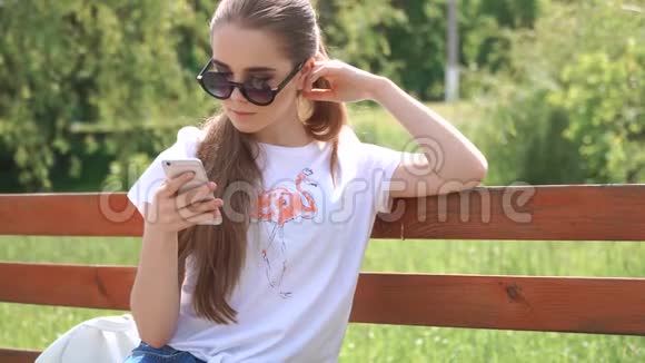 女孩在智能手机上说话坐在一个公园里坐在一张绿树叶的木凳上视频的预览图