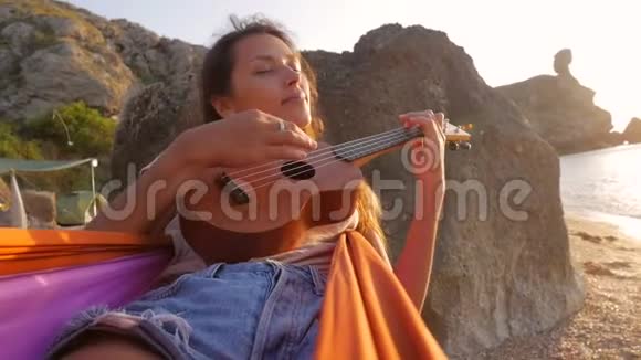 年轻迷人的嬉皮士混合种族女孩在吊床上摇摆在海滩上弹四弦琴吉他慢动作视频的预览图