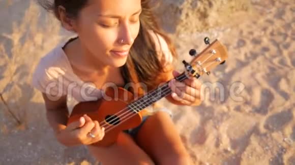 美丽的混血女人坐在沙滩上弹着四弦琴吉他高清慢镜头视频的预览图