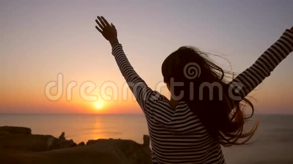 年轻的混血白种人女孩站在悬崖边张开双臂迎接海面上令人惊叹的日落视频的预览图