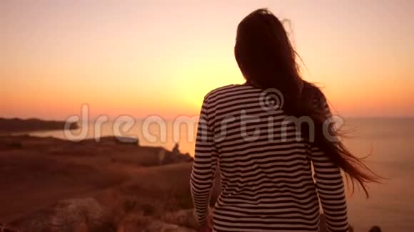 年轻的混血白种人女孩走在悬崖的边缘张开双臂迎接令人惊叹的近海日落视频的预览图