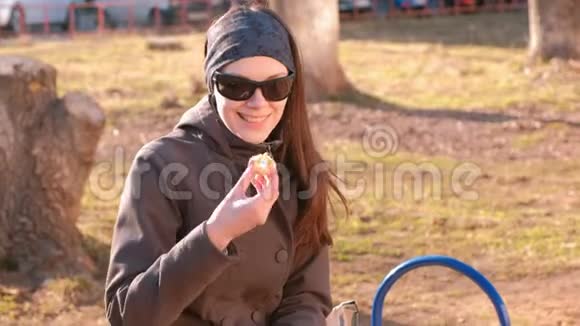 年轻的黑发女人戴着太阳镜在公园的长凳上吃橘子视频的预览图