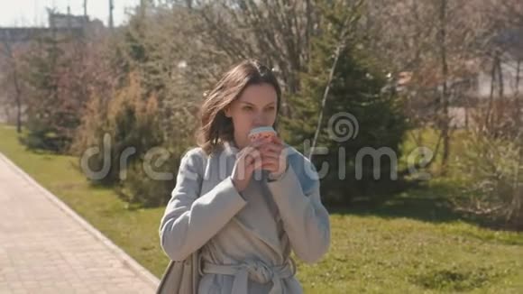 年轻的黑发女人穿着外套在城市公园散步喝咖啡春天视频的预览图