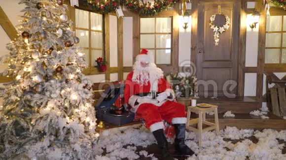 圣诞老人坐在房子附近的圣诞树之间喝牛奶吃饼干听乙烯基的圣诞歌曲视频的预览图