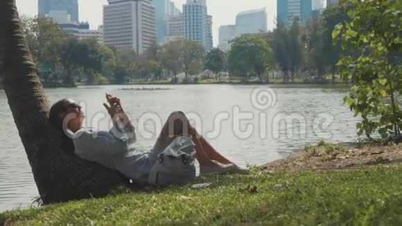 在公园里用手机躺在棕榈树下的年轻女子视频的预览图