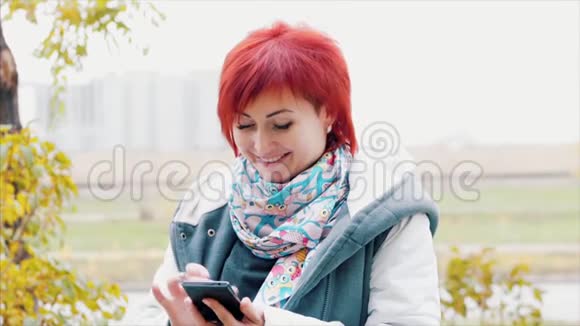 秋天漂亮的女人有着白种人的眼睛有着炽热的头发在街上兴高采烈地使用手机视频的预览图