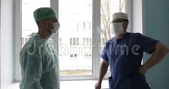 两名穿着制服和口罩的年轻医生正对着窗户在医院里交谈视频的预览图