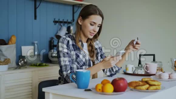 微笑的女人在家里的厨房里吃早餐时用智能手机和信用卡在网上购物视频的预览图