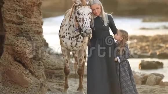 妈妈和女儿穿着优雅的衣服在阳光明媚的阳光下带着美丽的斑点马走在岩石海岸上视频的预览图