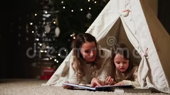 母亲和女儿穿着白色的毛衣读了一本童话故事和她躺在一个帐篷里视频的预览图