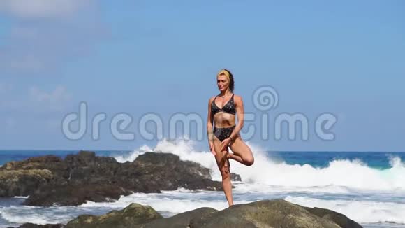 穿比基尼的年轻女孩一条腿站着做瑜伽站在海滩上的岩石上沙滩上有黑色的沙子视频的预览图