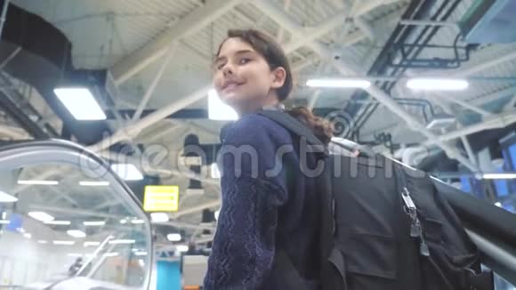 一个年轻的旅游女孩带着背包提着手提箱沿着自动扶梯上了机场等待飞机的生活方式视频的预览图