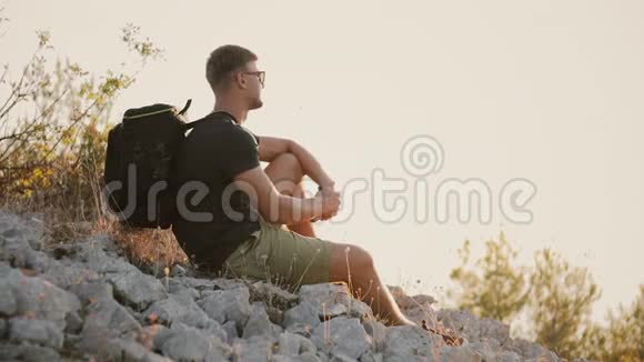 一位带着一个大背包的游客坐在岩石上在日落时休息视频的预览图